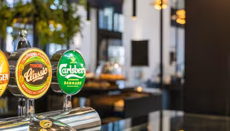 Nahaufnahme von Bierhähnen mit den Aufschriften Tuborg Classic und Carlsberg in einer Bar. Der Hintergrund ist modern eingerichtet und mit Pflanzen, Lichtern und unscharfen Möbeln gestaltet, was für eine gemütliche und einladende Atmosphäre sorgt.
