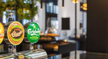Nahaufnahme von Bierhähnen mit den Aufschriften Tuborg Classic und Carlsberg in einer Bar. Der Hintergrund ist modern eingerichtet und mit Pflanzen, Lichtern und unscharfen Möbeln gestaltet, was für eine gemütliche und einladende Atmosphäre sorgt.