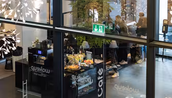 Blick durch die Glasfenster eines modernen Cafés mit einer Theke, an der Snacks und Getränke angeboten werden. Drinnen stehen Menschen an der Theke, darüber ist ein grünes Ausgangsschild zu sehen. Moderne Beleuchtung und Dekorationselemente sind zu sehen.