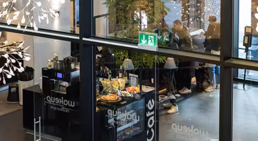 Blick durch die Glasfenster eines modernen Cafés mit einer Theke, an der Snacks und Getränke angeboten werden. Drinnen stehen Menschen an der Theke, darüber ist ein grünes Ausgangsschild zu sehen. Moderne Beleuchtung und Dekorationselemente sind zu sehen.