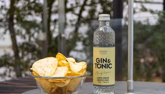 Eine Schüssel Kartoffelchips steht neben einer durchsichtigen Flasche mit der Aufschrift „Gin & Tonic“ auf einem Tisch im Freien. Im Hintergrund ist verschwommenes Grün zu sehen, das an eine Gartenkulisse erinnert.