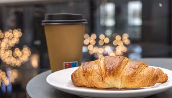 Ein butteriges Croissant liegt auf einem weißen Teller neben einem braunen Kaffeebecher zum Mitnehmen. Der Hintergrund ist sanft durch leuchtende Bokeh-Lichter verwischt und schafft so eine gemütliche Café-Atmosphäre.