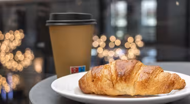 Ein butteriges Croissant liegt auf einem weißen Teller neben einem braunen Kaffeebecher zum Mitnehmen. Der Hintergrund ist sanft durch leuchtende Bokeh-Lichter verwischt und schafft so eine gemütliche Café-Atmosphäre.