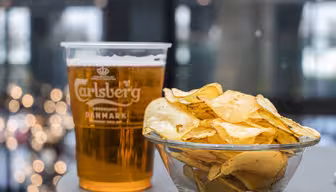 Ein durchsichtiger Plastikbecher mit Carlsberg-Bier steht neben einer Glasschale mit Kartoffelchips. Der Hintergrund ist durch sanftes Licht verwischt und schafft so eine gemütliche Atmosphäre.