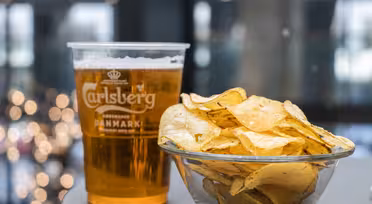 Ein durchsichtiger Plastikbecher mit Carlsberg-Bier steht neben einer Glasschale mit Kartoffelchips. Der Hintergrund ist durch sanftes Licht verwischt und schafft so eine gemütliche Atmosphäre.