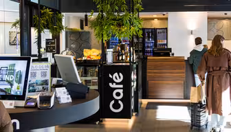 Ein modernes Café-Interieur mit heller und einladender Atmosphäre. Die Theke ist mit Gebäck und Getränken gefüllt. Im Vordergrund steht ein Touchscreen-Kiosk. Zwei Personen stehen in der Schlange, Pflanzen hängen als Dekoration von der Decke.