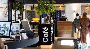 Ein modernes Café-Interieur mit heller und einladender Atmosphäre. Die Theke ist mit Gebäck und Getränken gefüllt. Im Vordergrund steht ein Touchscreen-Kiosk. Zwei Personen stehen in der Schlange, Pflanzen hängen als Dekoration von der Decke.