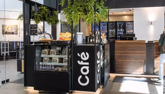 Eine moderne Café-Theke mit einer Glasvitrine mit Gebäck, Flaschen und abgepackten Waren. Ein großes Schild mit der Aufschrift „Café“ ist sichtbar. Darüber hängen Grünpflanzen, dahinter befindet sich eine Holztheke mit Kühltheke. Heller, geräumiger Innenraum.