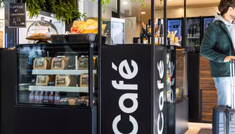 Ein modernes Café in Schwarz-Weiß präsentiert Snacks, Kaffee und Wein. Ein Mann in grüner Jacke steht mit einem Koffer da und schaut weg. Auf einem Schild steht „Café“, und ein kleineres Schild wirbt für Kaffee und Croissant für 4,9 Einheiten.