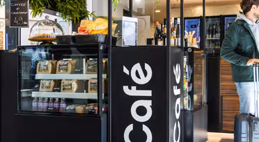 Ein modernes Café in Schwarz-Weiß präsentiert Snacks, Kaffee und Wein. Ein Mann in grüner Jacke steht mit einem Koffer da und schaut weg. Auf einem Schild steht „Café“, und ein kleineres Schild wirbt für Kaffee und Croissant für 4,9 Einheiten.