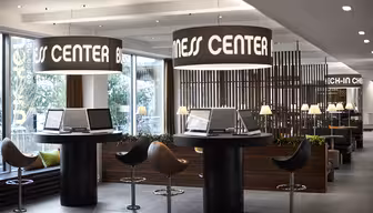 Modernes Business Center mit Computerarbeitsplätzen und Hochstühlen. Große zylindrische Leuchten hängen darüber und zeigen den Schriftzug „BUSINESS CENTER“. Im Hintergrund befinden sich ein Check-in-Schalter und sanft beleuchtete Lampen, die eine professionelle und einladende Atmosphäre schaffen.