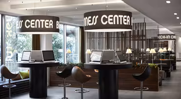 Modernes Business Center mit Computerarbeitsplätzen und Hochstühlen. Große zylindrische Leuchten hängen darüber und zeigen den Schriftzug „BUSINESS CENTER“. Im Hintergrund befinden sich ein Check-in-Schalter und sanft beleuchtete Lampen, die eine professionelle und einladende Atmosphäre schaffen.