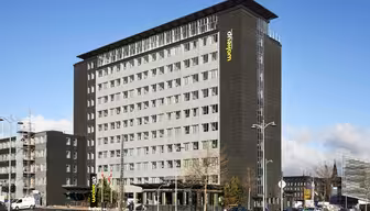 Ein modernes, mehrstöckiges Hotelgebäude mit dunkler Fassade und großen rechteckigen Fenstern. Der Name „Wakeup“ prangt in Grün und Gelb an der Fassade. Das Gebäude ist von einigen Bäumen, parkenden Autos und einem strahlend blauen Himmel umgeben.
