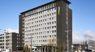 Ein modernes, mehrstöckiges Hotelgebäude mit dunkler Fassade und großen rechteckigen Fenstern. Der Name „Wakeup“ prangt in Grün und Gelb an der Fassade. Das Gebäude ist von einigen Bäumen, parkenden Autos und einem strahlend blauen Himmel umgeben.