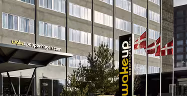 Modernes Hotelgebäude mit Glasfassade und dem Schild „wakeupcopenhagen.com“. Neben dem Eingang wehen drei dänische Flaggen. Die Architektur zeichnet sich durch klare Linien und große Fenster aus. Im Vordergrund sind Büsche und Wege zu sehen.