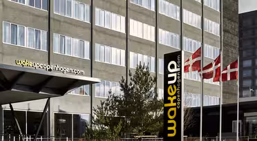 Modernes Hotelgebäude mit Glasfassade und dem Schild „wakeupcopenhagen.com“. Neben dem Eingang wehen drei dänische Flaggen. Die Architektur zeichnet sich durch klare Linien und große Fenster aus. Im Vordergrund sind Büsche und Wege zu sehen.