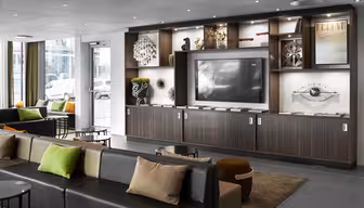 Moderne Hotellobby mit zeitgenössischem Dekor. Verfügt über eine große Schrankwand mit Fernseher, Kunstwerken und Dekoartikeln. Zu den Sitzgelegenheiten gehören stilvolle Sofas und Ottomanen mit bunten Kissen. Große Fenster lassen Tageslicht herein.
