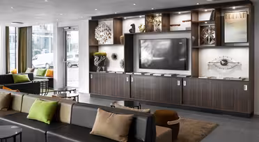 Moderne Hotellobby mit zeitgenössischem Dekor. Verfügt über eine große Schrankwand mit Fernseher, Kunstwerken und Dekoartikeln. Zu den Sitzgelegenheiten gehören stilvolle Sofas und Ottomanen mit bunten Kissen. Große Fenster lassen Tageslicht herein.