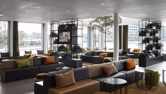 Moderne Hotellobby mit großen Fenstern, dunklen Sofas mit orangefarbenen und beigen Kissen. Grüne Pflanzen werden auf Regalen präsentiert und überall stehen runde Glastische verteilt. Natürliches Licht erhellt den Raum und schafft eine einladende Atmosphäre.