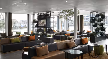 Moderne Hotellobby mit großen Fenstern, dunklen Sofas mit orangefarbenen und beigen Kissen. Grüne Pflanzen werden auf Regalen präsentiert und überall stehen runde Glastische verteilt. Natürliches Licht erhellt den Raum und schafft eine einladende Atmosphäre.