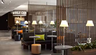 Eine moderne Wellness-Lounge mit Holzlamellen, die die Sitzbereiche voneinander trennen. Schwarze Sofas mit bunten Kissen stehen neben runden Tischen und Lampen. Grüne Pflanzen sorgen für einen Hauch von Natur. Deckenleuchten erhellen den Raum.
