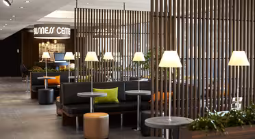 Eine moderne Wellness-Lounge mit Holzlamellen, die die Sitzbereiche voneinander trennen. Schwarze Sofas mit bunten Kissen stehen neben runden Tischen und Lampen. Grüne Pflanzen sorgen für einen Hauch von Natur. Deckenleuchten erhellen den Raum.