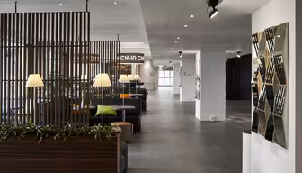 Moderner Lobbybereich mit eleganten Holztrennwänden und Grünpflanzen. Sanftes Licht von Tischlampen erhellt die Sitzbereiche mit grünen und orangefarbenen Kissen. Rechts befindet sich ein geometrischer Spiegelwand-Akzent. Der Boden ist gefliest, der Raum offen und hell.