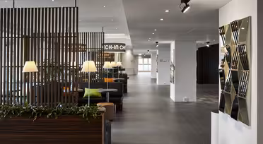 Moderner Lobbybereich mit eleganten Holztrennwänden und Grünpflanzen. Sanftes Licht von Tischlampen erhellt die Sitzbereiche mit grünen und orangefarbenen Kissen. Rechts befindet sich ein geometrischer Spiegelwand-Akzent. Der Boden ist gefliest, der Raum offen und hell.