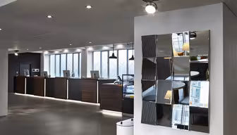 Moderne Hotelrezeption mit einer Reihe dunkler Holztische und Computer vor einer Wand mit großen Fenstern. An der rechten Wand ist ein dekorativer, mehrteiliger Spiegel angebracht, der Teile des Raumes reflektiert. Der Boden ist mit grauen Fliesen ausgelegt.