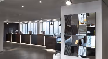 Moderne Hotelrezeption mit einer Reihe dunkler Holztische und Computer vor einer Wand mit großen Fenstern. An der rechten Wand ist ein dekorativer, mehrteiliger Spiegel angebracht, der Teile des Raumes reflektiert. Der Boden ist mit grauen Fliesen ausgelegt.