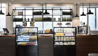 Ein modernes Café-Interieur mit zwei Glasvitrinen für Getränke, Salate und Snacks. Darüber befinden sich Regale mit Weinflaschen und Pflanzen. Von der Decke hängen Pendelleuchten, und auf der linken Theke steht eine Registrierkasse.