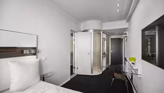 Modernes Hotelzimmer mit einem sauber gemachten Bett, einer verglasten Dusche und eleganten Möbeln. Die Wände sind weiß, und gegenüber dem Bett befindet sich ein Flachbildfernseher. Das Zimmer ist minimalistisch gestaltet und sanft beleuchtet.