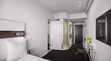 Modernes Hotelzimmer mit einem sauber gemachten Bett, einer verglasten Dusche und eleganten Möbeln. Die Wände sind weiß, und gegenüber dem Bett befindet sich ein Flachbildfernseher. Das Zimmer ist minimalistisch gestaltet und sanft beleuchtet.