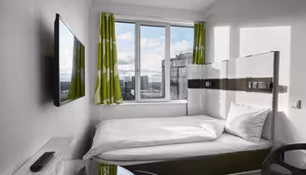 Ein kleines, modernes Hotelzimmer verfügt über ein ordentlich gemachtes Bett unter einem großen Fenster mit grün gemusterten Vorhängen. An der Wand hängt ein Flachbildfernseher, im Vordergrund steht ein Glastisch mit Stuhl. Der Blick nach draußen zeigt Gebäude und einen bewölkten Himmel.