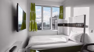 Ein kleines, modernes Hotelzimmer verfügt über ein ordentlich gemachtes Bett unter einem großen Fenster mit grün gemusterten Vorhängen. An der Wand hängt ein Flachbildfernseher, im Vordergrund steht ein Glastisch mit Stuhl. Der Blick nach draußen zeigt Gebäude und einen bewölkten Himmel.
