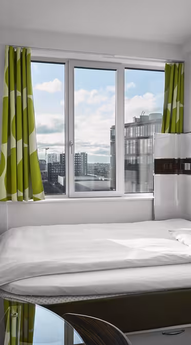 Ein kleines, modernes Hotelzimmer verfügt über ein ordentlich gemachtes Bett unter einem großen Fenster mit grün gemusterten Vorhängen. An der Wand hängt ein Flachbildfernseher, im Vordergrund steht ein Glastisch mit Stuhl. Der Blick nach draußen zeigt Gebäude und einen bewölkten Himmel.