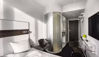 Modernes Hotelzimmer mit Bett, transparenter Glasdusche, schwarzem Stuhl und elegantem Schreibtisch mit Fernseher an der Wand. Weiße Wände und dunkler Teppich sorgen für einen minimalistischen Look. Ein grüner Gegenstand steht auf dem Schreibtisch neben dem Wandtelefon.