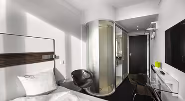 Modernes Hotelzimmer mit Bett, transparenter Glasdusche, schwarzem Stuhl und elegantem Schreibtisch mit Fernseher an der Wand. Weiße Wände und dunkler Teppich sorgen für einen minimalistischen Look. Ein grüner Gegenstand steht auf dem Schreibtisch neben dem Wandtelefon.