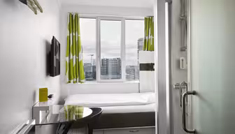 Kompaktes Hotelzimmer mit Einzelbett, grün-weißen Vorhängen, Flachbildfernseher und Glasduschtür. Schreibtisch und Stuhl stehen am Fenster und bieten einen Blick auf die Stadtgebäude im Hintergrund.