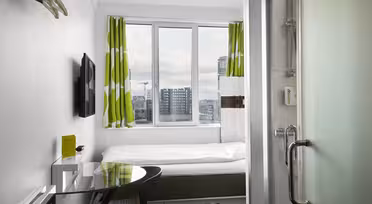 Kompaktes Hotelzimmer mit Einzelbett, grün-weißen Vorhängen, Flachbildfernseher und Glasduschtür. Schreibtisch und Stuhl stehen am Fenster und bieten einen Blick auf die Stadtgebäude im Hintergrund.