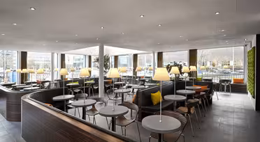 Ein modernes, geräumiges Café mit runden Tischen und Holzstühlen. Sanftes Licht von Tischlampen sorgt für eine gemütliche Atmosphäre. Große Fenster lassen Tageslicht herein, und rechts befindet sich eine grüne Wand. Der Raum ist elegant und modern gestaltet.