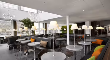 Ein modernes Café mit runden Tischen und schwarzen Sitzpolstern. Jeder Tisch verfügt über eine Lampe mit weißem Schirm. Die Sitze sind mit orangefarbenen und grünen Kissen ausgestattet. Große Fenster und dekorative Pflanzen schaffen eine helle, offene Atmosphäre.