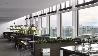 Moderne Lounge mit großen Fenstern und Blick auf die Stadt. Sie verfügt über Reihen von Tischen und Stühlen, Grünpflanzen und einen Buffetbereich mit Metalltabletts. Deckenbeleuchtung und ein klares, modernes Design schaffen eine einladende Atmosphäre.