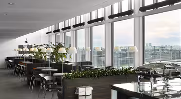 Moderne Lounge mit großen Fenstern und Blick auf die Stadt. Sie verfügt über Reihen von Tischen und Stühlen, Grünpflanzen und einen Buffetbereich mit Metalltabletts. Deckenbeleuchtung und ein klares, modernes Design schaffen eine einladende Atmosphäre.