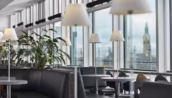 Moderner Wartebereich am Flughafen mit stilvollen schwarzen Sitzgelegenheiten und Tischen unter eleganten weißen Lampen. Große Fenster bieten den Blick auf einen fernen Turm vor bewölktem Himmel. Grüne Pflanzen verleihen dem modernen Ambiente einen Hauch von Natur.