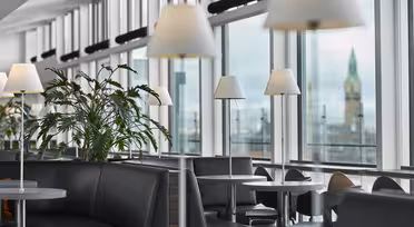 Moderner Wartebereich am Flughafen mit stilvollen schwarzen Sitzgelegenheiten und Tischen unter eleganten weißen Lampen. Große Fenster bieten den Blick auf einen fernen Turm vor bewölktem Himmel. Grüne Pflanzen verleihen dem modernen Ambiente einen Hauch von Natur.