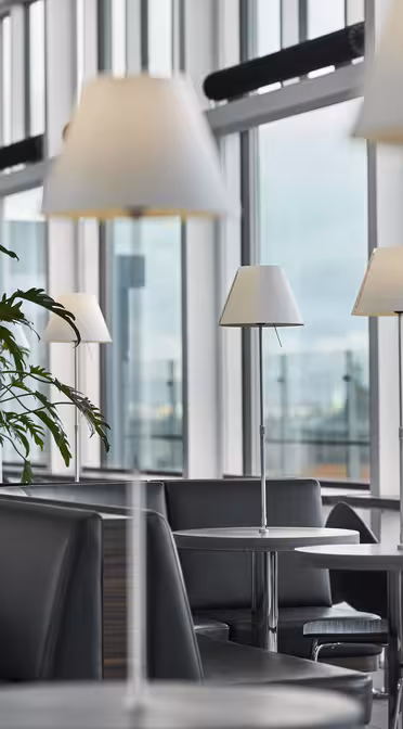 Moderner Wartebereich am Flughafen mit stilvollen schwarzen Sitzgelegenheiten und Tischen unter eleganten weißen Lampen. Große Fenster bieten den Blick auf einen fernen Turm vor bewölktem Himmel. Grüne Pflanzen verleihen dem modernen Ambiente einen Hauch von Natur.