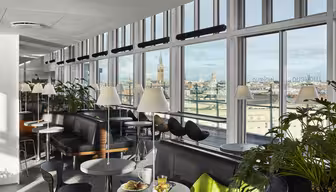 Eine moderne Lounge mit großen Fenstern und Blick auf die Stadt. Der Raum ist mit Sofas, schwarzen Stühlen und kleinen runden Tischen mit Lampen ausgestattet. An einigen Tischen stehen Speisen und Getränke bereit. Topfpflanzen schmücken den Bereich und schaffen eine gemütliche und einladende Atmosphäre.