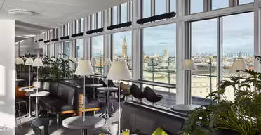 Eine moderne Lounge mit großen Fenstern und Blick auf die Stadt. Der Raum ist mit Sofas, schwarzen Stühlen und kleinen runden Tischen mit Lampen ausgestattet. An einigen Tischen stehen Speisen und Getränke bereit. Topfpflanzen schmücken den Bereich und schaffen eine gemütliche und einladende Atmosphäre.