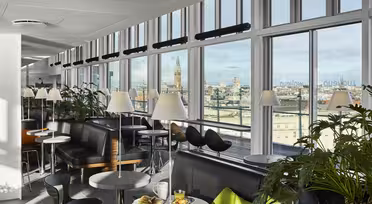 Eine moderne Lounge mit großen Fenstern und Blick auf die Stadt. Der Raum ist mit Sofas, schwarzen Stühlen und kleinen runden Tischen mit Lampen ausgestattet. An einigen Tischen stehen Speisen und Getränke bereit. Topfpflanzen schmücken den Bereich und schaffen eine gemütliche und einladende Atmosphäre.